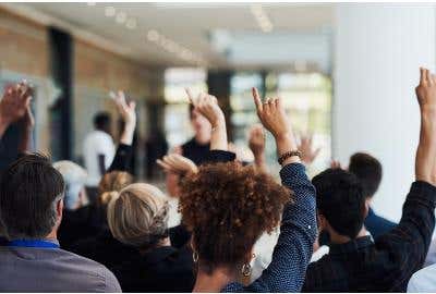 Classroom full of people, many of whom are raising their hands