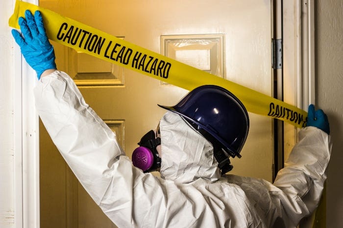 Worker in hazard protection gear putting up caution tape which reads "Caution Lead Hazard"